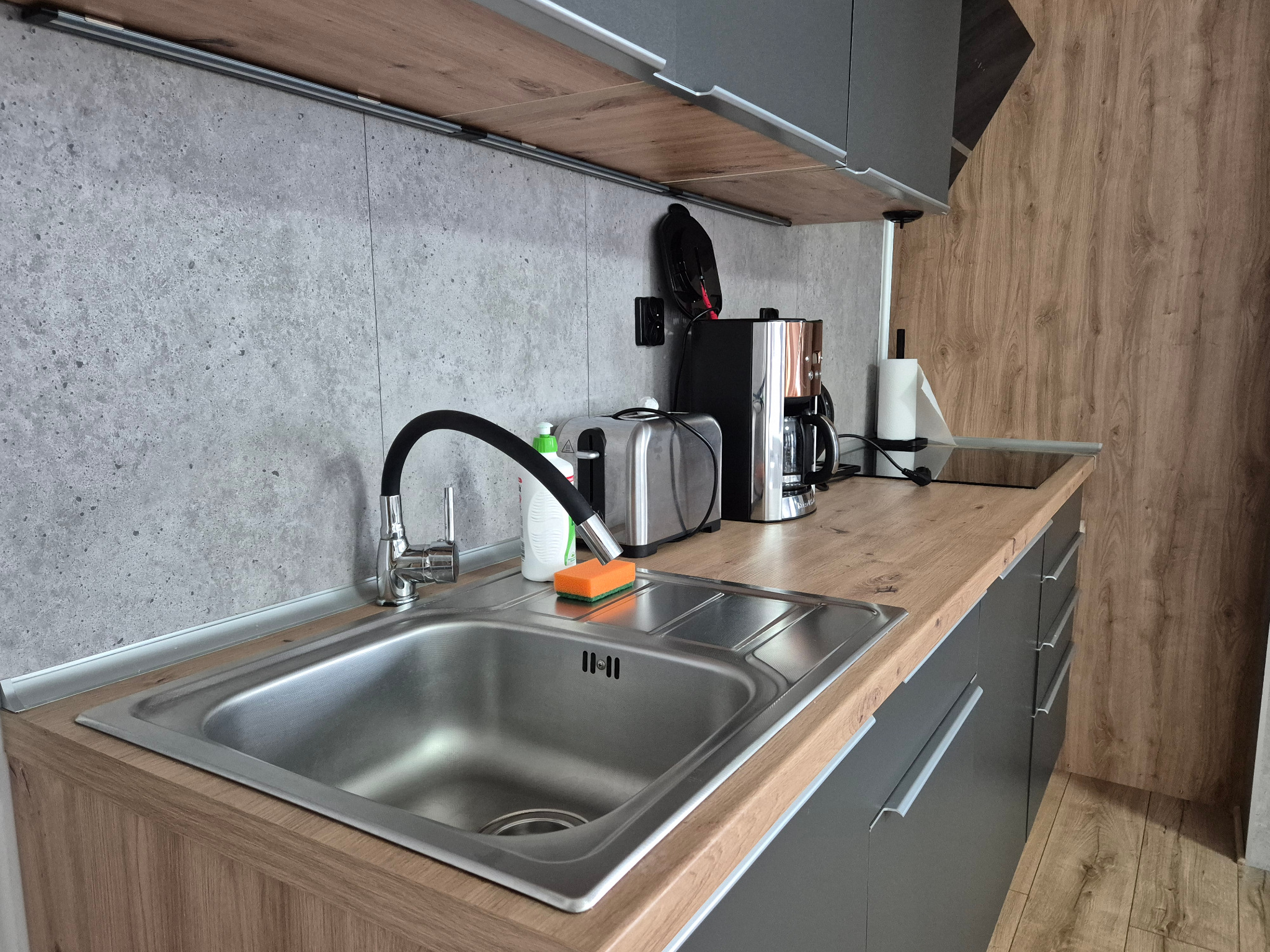 Nowoczesna kuchnia: metalowy zlew, bateria z elastyczną wylewką, gąbka i płyn do naczyń na tle blatu z drewnianym wzorem i szarych szafek.