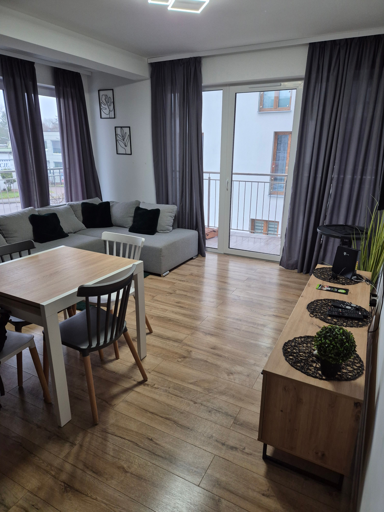 Jasne wnętrze mieszkania z drewnianą podłogą, szarą sofą i stołem z krzesłami. Widok na balkon i okna z szarymi zasłonami. Minimalistyczne dekoracje ścienne.