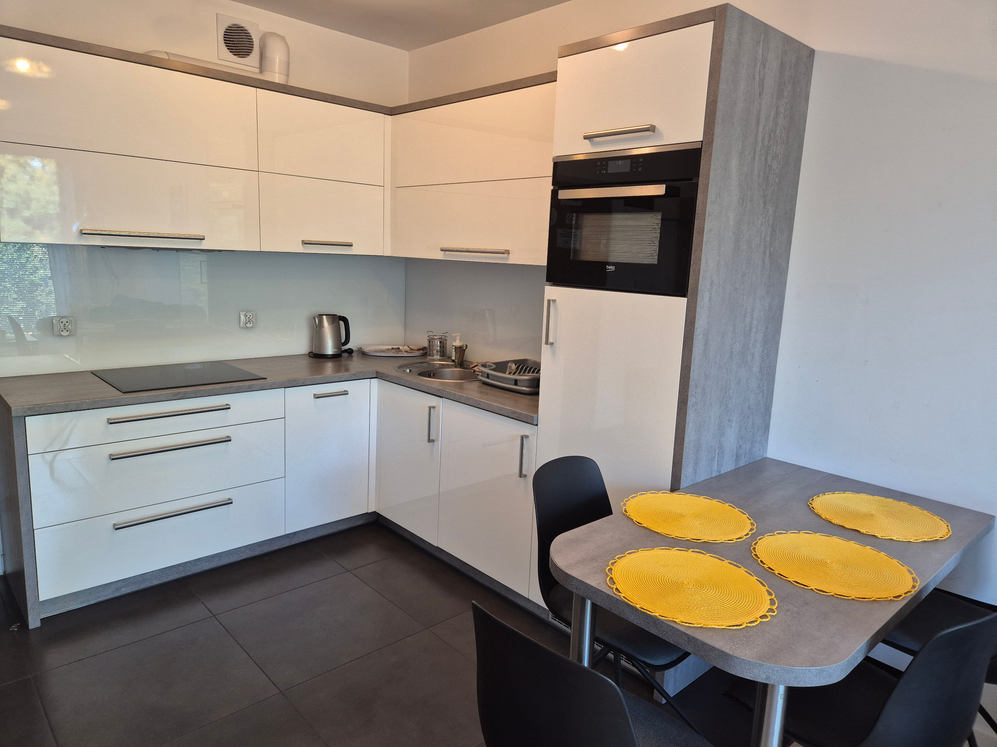 Błyszcząca, biała kuchnia w nowoczesnym stylu z szarym blatem i czarną płytą indukcyjną. Na stole cztery żółte podkładki. Minimalistyczny design.