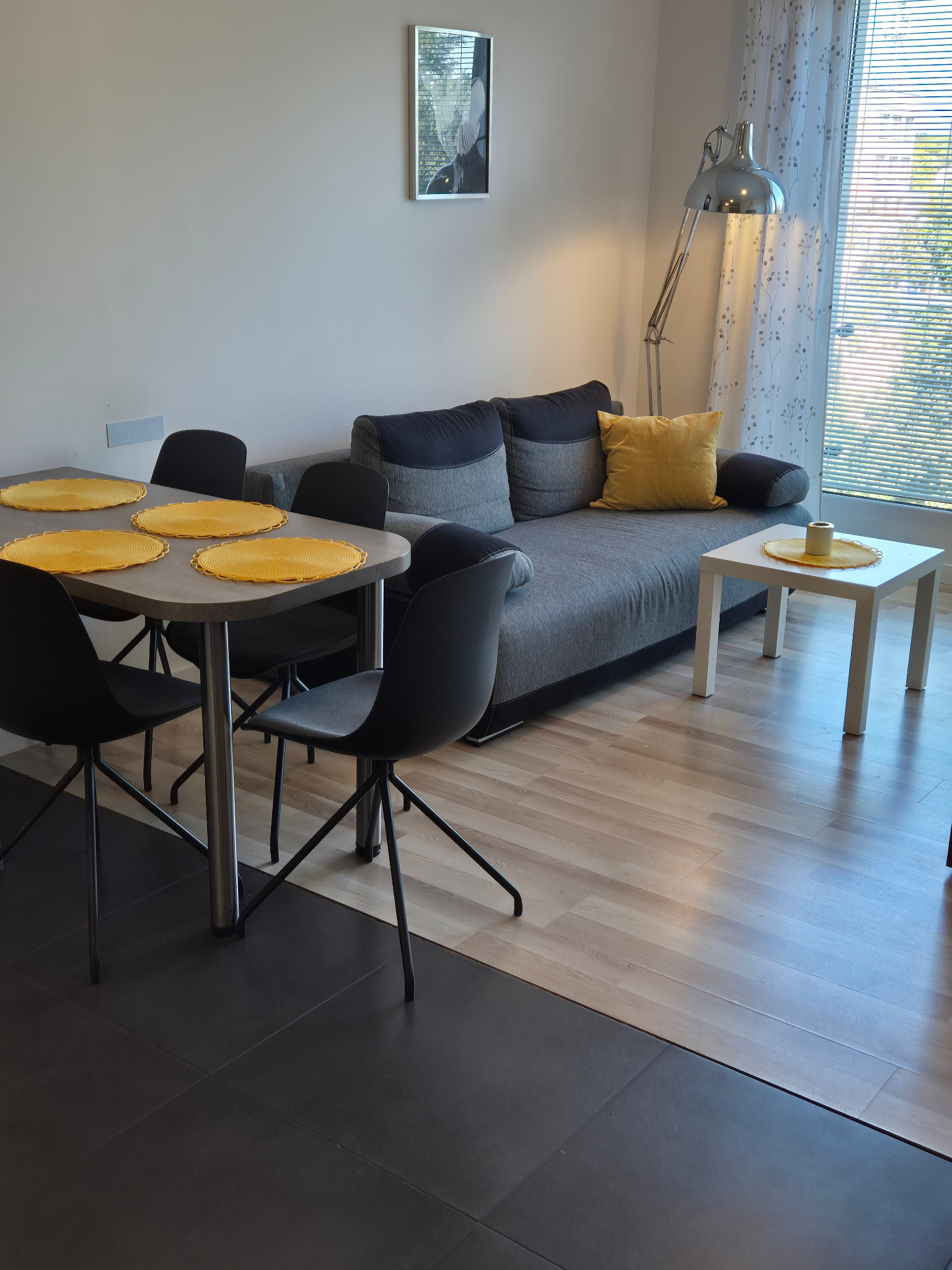 Jasne wnętrze pokoju dziennego: stół z żółtymi podkładkami, szara sofa z poduszką, lampa podłogowa i okno z żaluzjami, tworzące przytulną atmosferę.