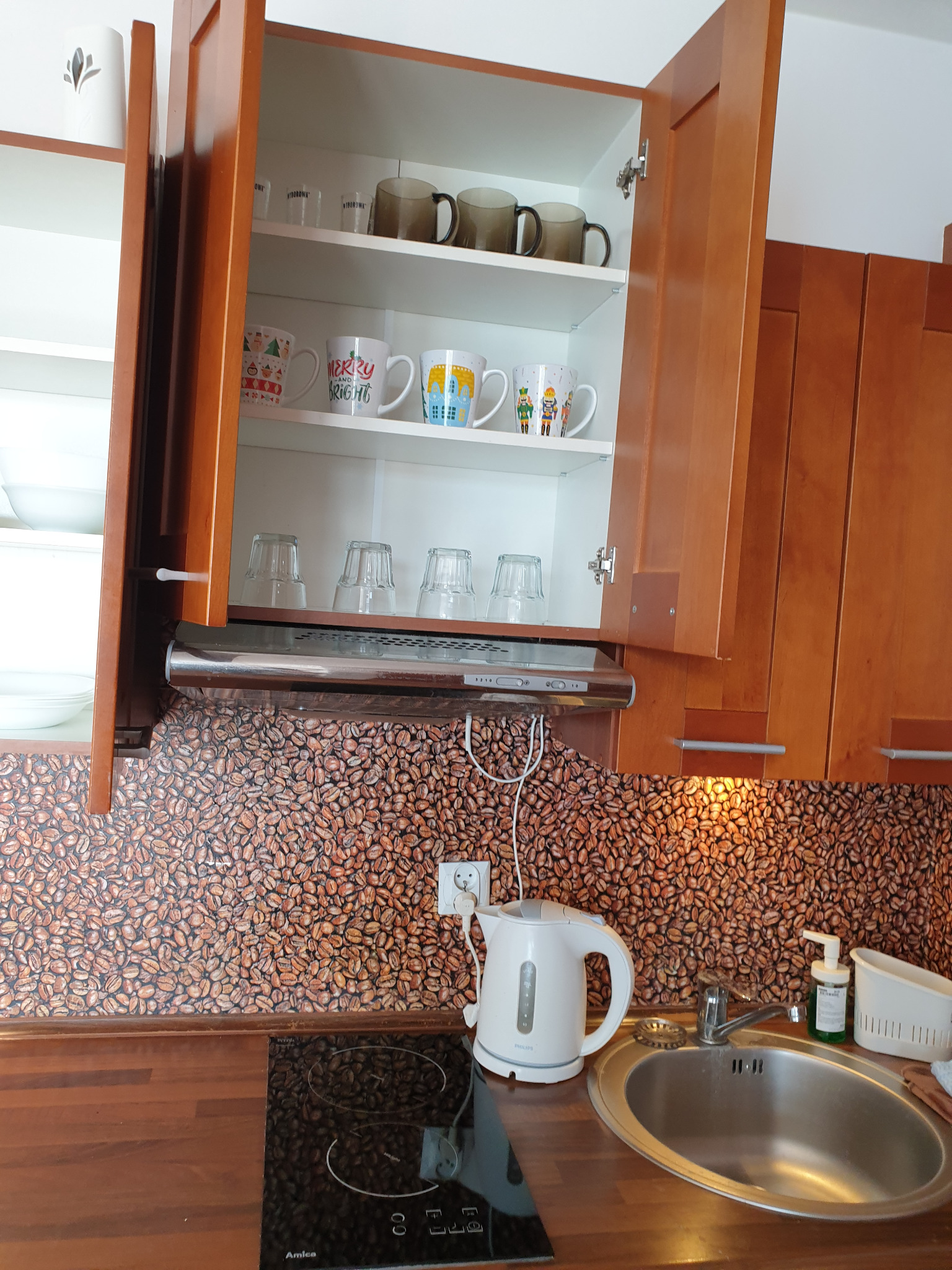 Umyta kuchnia: błyszczący zlew, czajnik i szafki z naczyniami na tle ściany z motywem ziaren kawy. Widoczne detale wyposażenia.