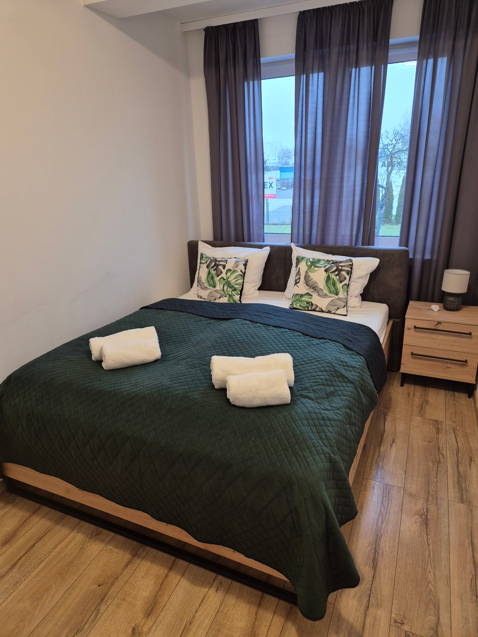 Przytulna sypialnia z łóżkiem, zieloną narzutą, białymi ręcznikami i poduszkami w tropikalne liście. Drewniana podłoga i zasłony w oknie.