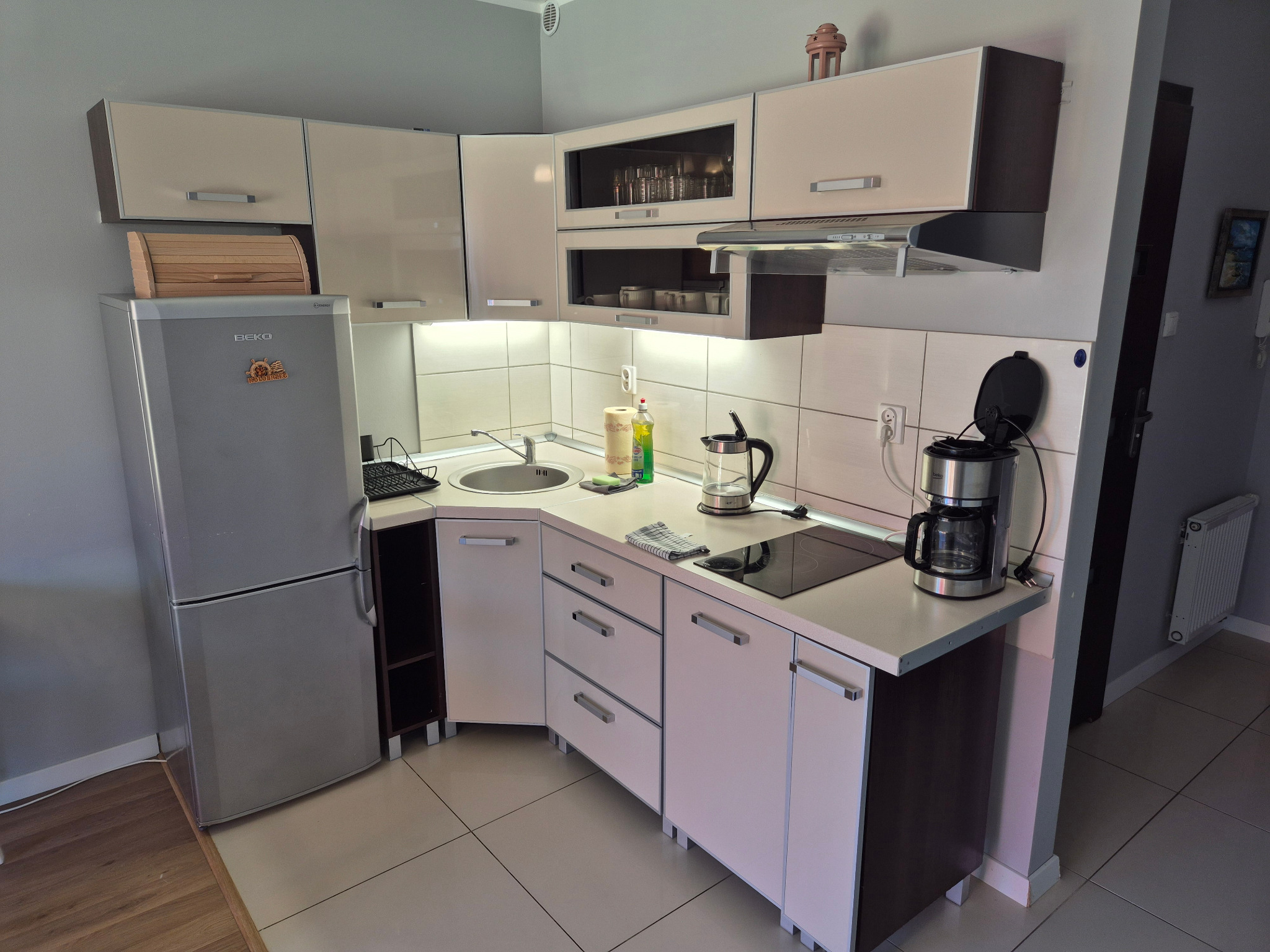 Jasna, nowoczesna kuchnia w aneksie z szarym sprzętem AGD, białymi płytkami i drewnianymi akcentami. Widok na blat z ekspresem do kawy i płytą grzewczą.