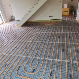 TERMECO TCHÓRZEWSCY SPÓŁKA JAWNA - Pomarańczowe rury ogrzewania podłogowego ułożone na czarnej macie z siatką w surowym wnętrzu domu, widok z góry na instalację przed wylaniem posadzki.