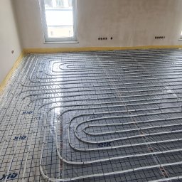 TERMECO TCHÓRZEWSCY SPÓŁKA JAWNA - Ułożone rury ogrzewania podłogowego na folii z siatką w surowym wnętrzu. Widoczne dwa okna, żółta taśma na ścianach. Instalacja przed wylaniem posadzki.