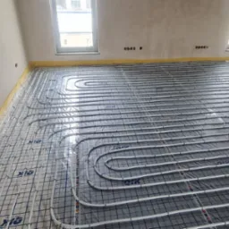 Ułożone rury ogrzewania podłogowego na folii z siatką w surowym wnętrzu. Widoczne dwa okna, żółta taśma na ścianach. Instalacja przed wylaniem posadzki.