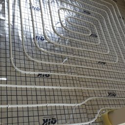 TERMECO TCHÓRZEWSCY SPÓŁKA JAWNA - Białe rury ogrzewania podłogowego ułożone na czarnej siatce z logo na folii izolacyjnej. Widok z góry, rury przymocowane plastikowymi klipsami, etap instalacji.