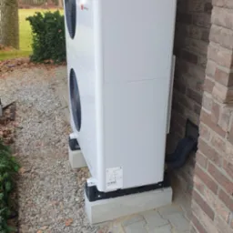 Biała pompa ciepła powietrze-woda, zamontowana na betonowych blokach przy ceglanym budynku, z widocznymi rurami wchodzącymi do ściany i otoczeniem z kamiennego żwiru.