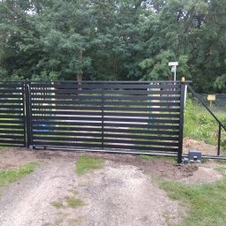 Spaw-Mat Mateusz Osiński - Nowoczesna, przesuwna brama wjazdowa w kolorze czarnym, z widocznym napędem elektrycznym i zębatką, na tle zieleni drzew i krzewów. Brama częściowo otwarta.