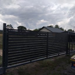Spaw-Mat Mateusz Osiński - Nowoczesna, przesuwna brama wjazdowa z poziomych profili, pomalowana na ciemny kolor, na tle zachmurzonego nieba i zieleni. Widoczny fragment ogrodzenia panelowego.