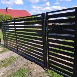 Spaw-Mat Mateusz Osiński - Nowoczesna, pozioma brama przesuwna w kolorze antracytu, częściowo otwarta. Widoczny dom z czerwoną dachówką i fragment osoby w tle, sugerujący montaż.