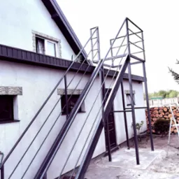 Metalowe schody zewnętrzne z balustradą, prowadzące do drzwi na piętrze domu. Widoczne drewno opałowe i drabina w tle, podkreślające funkcjonalność przestrzeni.