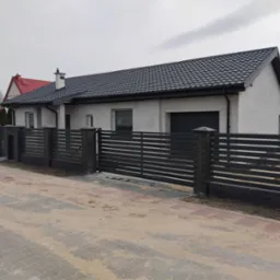 Nowoczesna brama wjazdowa z poziomych, metalowych profili, osadzona na murowanych słupkach z cegły klinkierowej, na tle domu z ciemną dachówką.