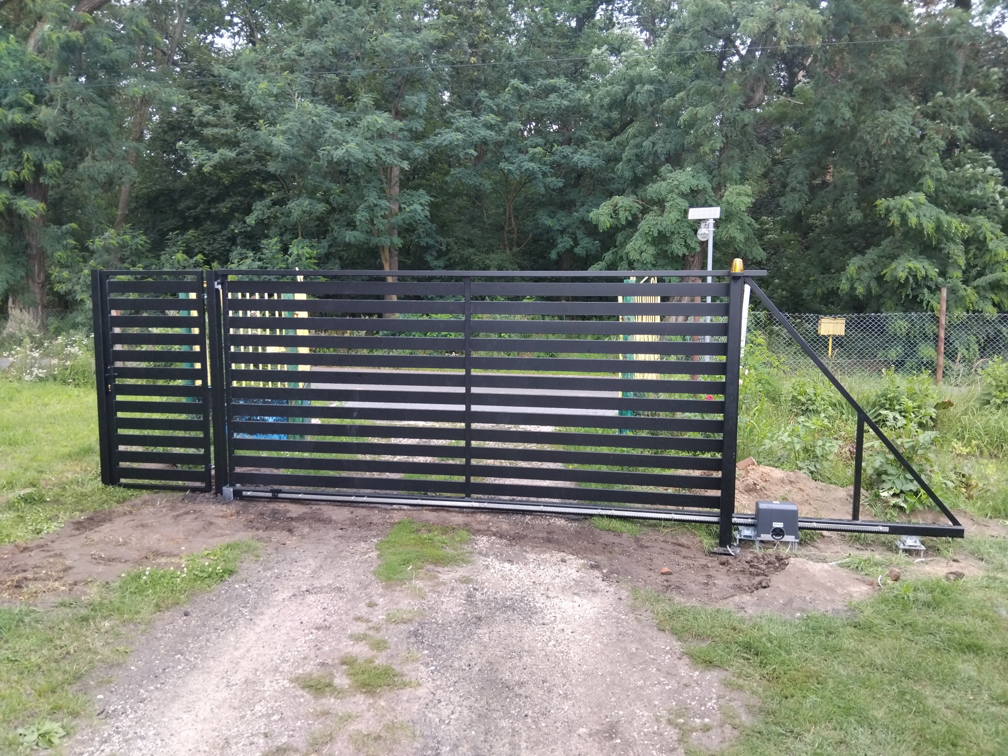 Nowoczesna, przesuwna brama wjazdowa w kolorze czarnym, z widocznym napędem elektrycznym i zębatką, na tle zieleni drzew i krzewów. Brama częściowo otwarta.