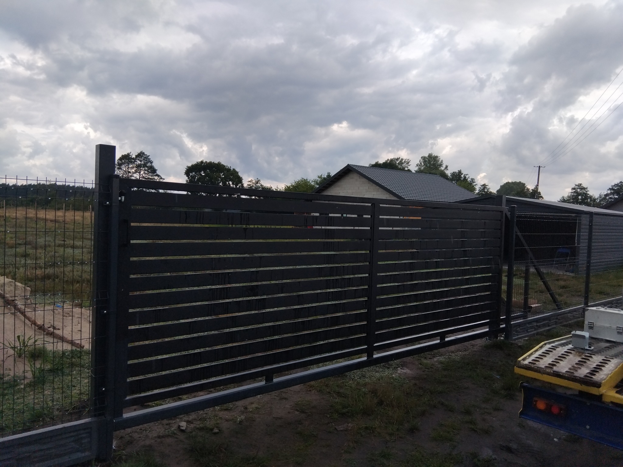 Nowoczesna, przesuwna brama wjazdowa z poziomych profili, pomalowana na ciemny kolor, na tle zachmurzonego nieba i zieleni. Widoczny fragment ogrodzenia panelowego.
