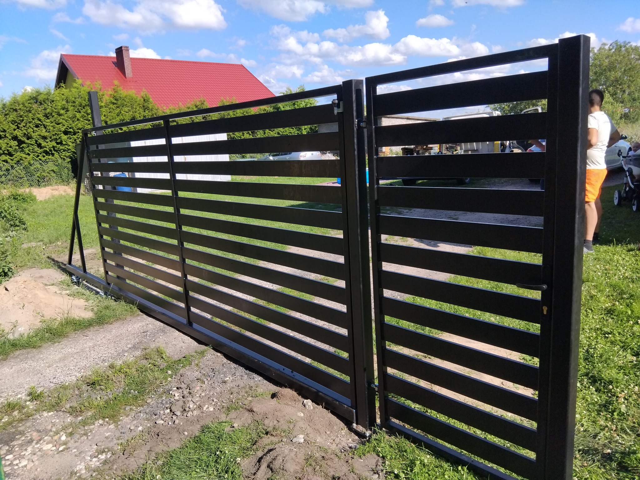 Nowoczesna, pozioma brama przesuwna w kolorze antracytu, częściowo otwarta. Widoczny dom z czerwoną dachówką i fragment osoby w tle, sugerujący montaż.