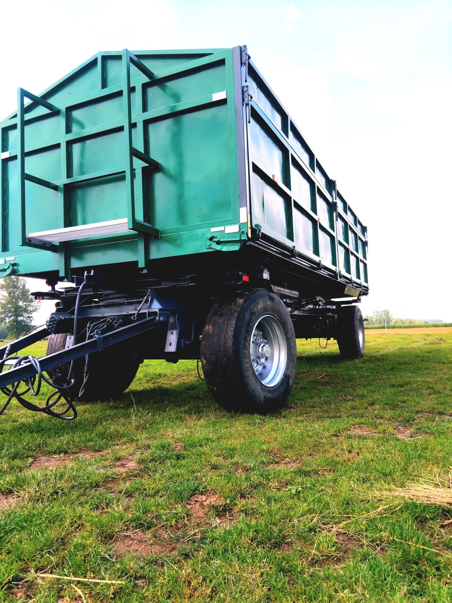Zielona przyczepa rolnicza z metalową konstrukcją na łące, widok z dołu, ukazujący detale podwozia i opony. Tło to jasne niebo i pole.