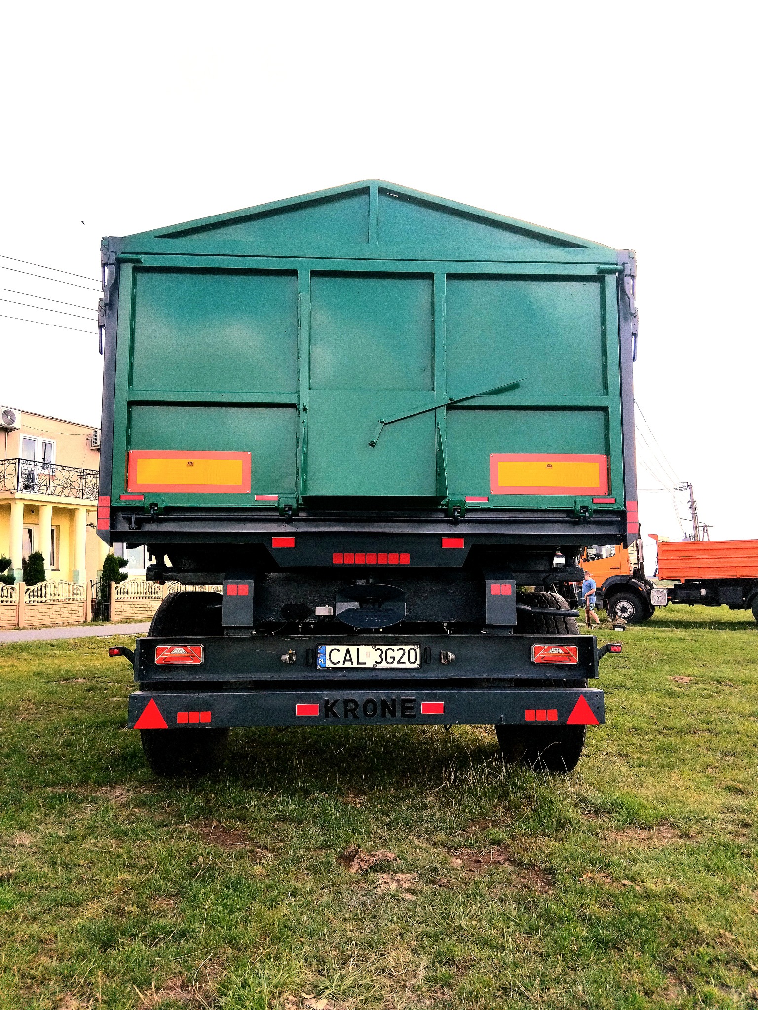 Zielona naczepa z tablicą rejestracyjną CAL 3G20 marki Krone, widziana z tyłu na trawiastym terenie, z fragmentem budynku i innymi pojazdami w tle.