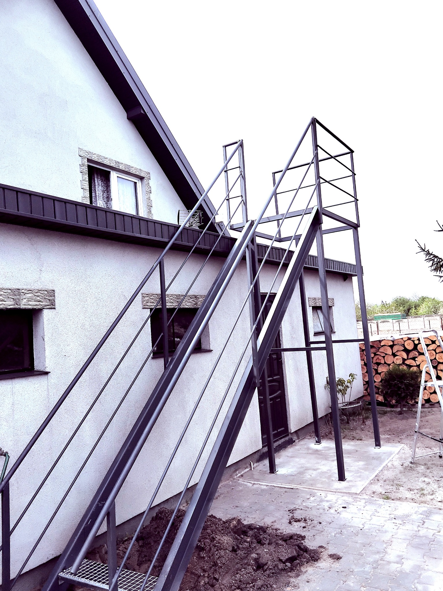 Metalowe schody zewnętrzne z balustradą, prowadzące do drzwi na piętrze domu. Widoczne drewno opałowe i drabina w tle, podkreślające funkcjonalność przestrzeni.