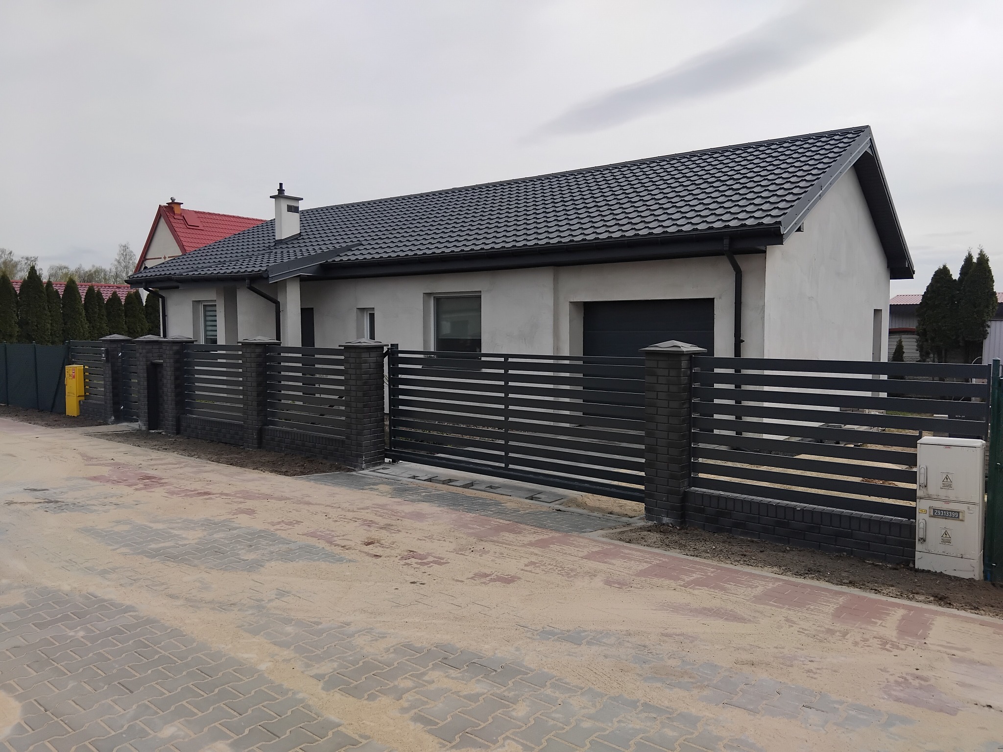 Nowoczesna brama wjazdowa z poziomych, metalowych profili, osadzona na murowanych słupkach z cegły klinkierowej, na tle domu z ciemną dachówką.