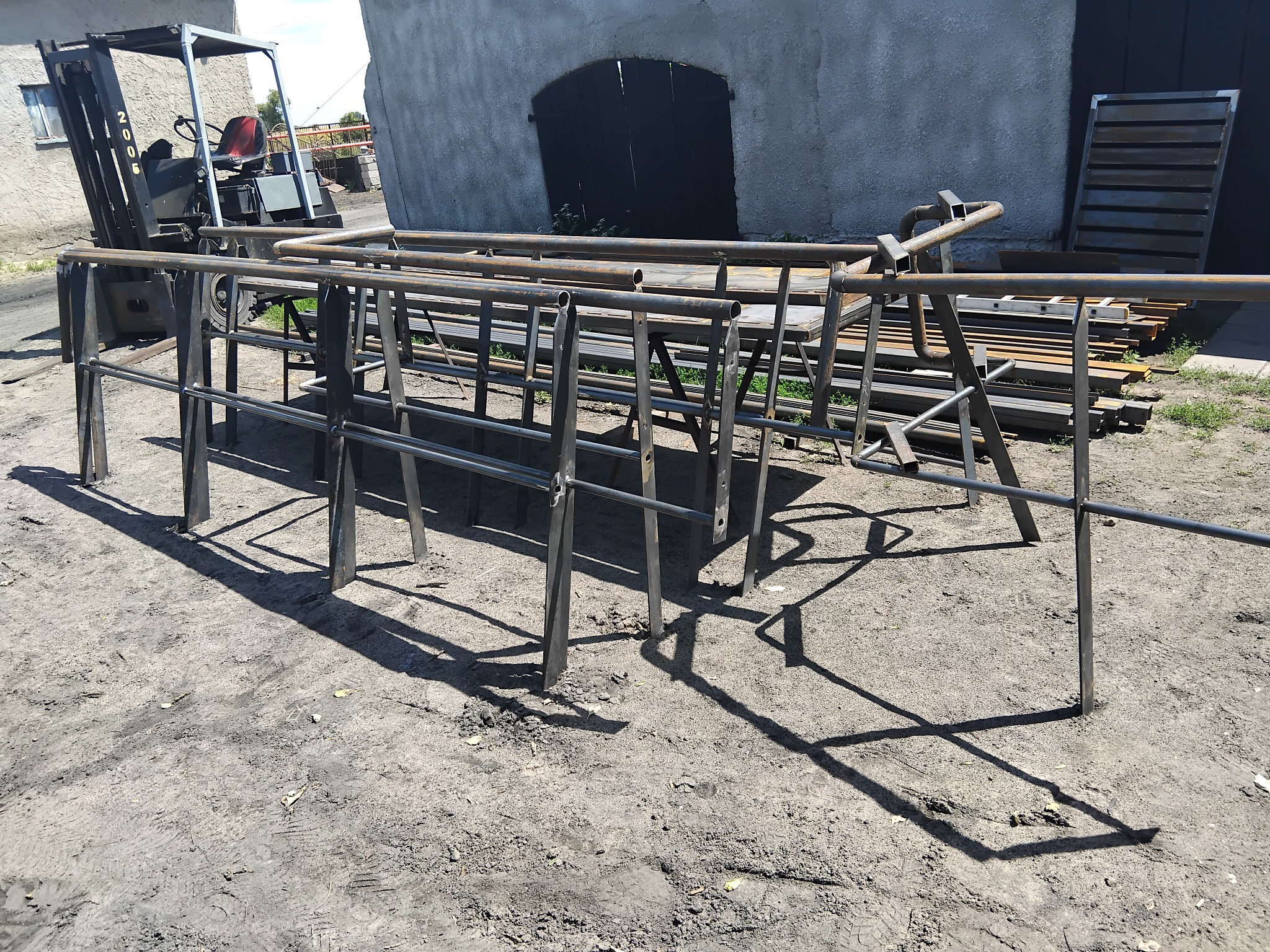 Surowe, metalowe elementy balustrady w warsztacie, częściowo oparte o wózek widłowy; w tle budynek z ciemnymi drzwiami i stos metalowych profili.