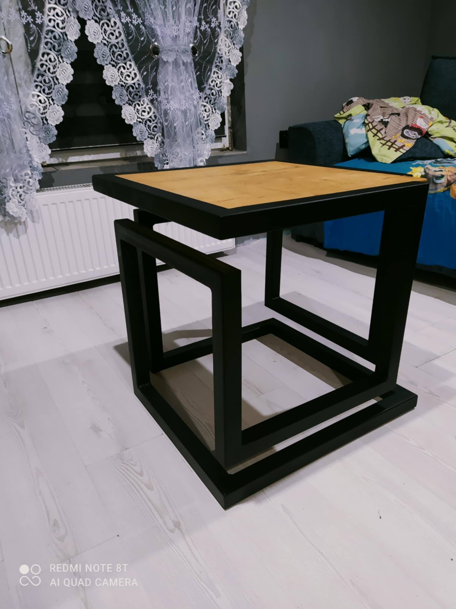 Nowoczesny, spawany stolik kawowy z czarną, stalową ramą i blatem z jasnego drewna. Geometryczna konstrukcja w pokoju.