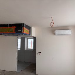 Majuko Sp. z o.o. - Montaż klimatyzacji kanałowej i ściennej w surowym wnętrzu. Widoczne zabezpieczenia folią i taśmą. Przewody elektryczne wystają ze ściany i sufitu.