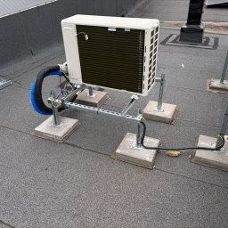 Majuko Sp. z o.o. - Zewnętrzna jednostka klimatyzacji na dachu, zamontowana na metalowej konstrukcji z betonowymi podstawami. Widoczne przewody i elementy instalacji.