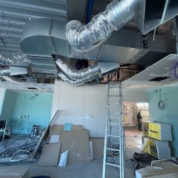 Majuko Sp. z o.o. - Montaż instalacji wentylacyjnej w surowym wnętrzu. Widoczne metalowe rury wentylacyjne pod sufitem, drabina i materiały budowlane. W tle osoba w roboczym ubraniu.