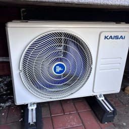 Majuko Sp. z o.o. - Biała jednostka zewnętrzna klimatyzacji Kaisai z niebieskim wentylatorem, zamontowana na czarnych wspornikach na czerwonej kostce brukowej, widok z dołu.