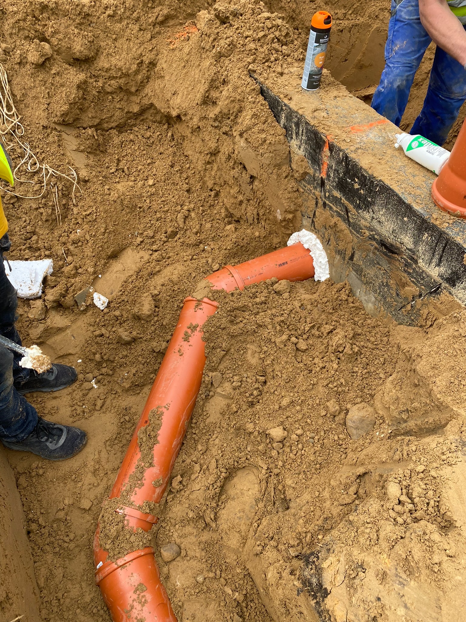 Pomarańczowa rura wentylacyjna w wykopie, zabezpieczona pianką montażową przy ścianie fundamentowej. Widoczne narzędzia i fragmenty odzieży pracowników.