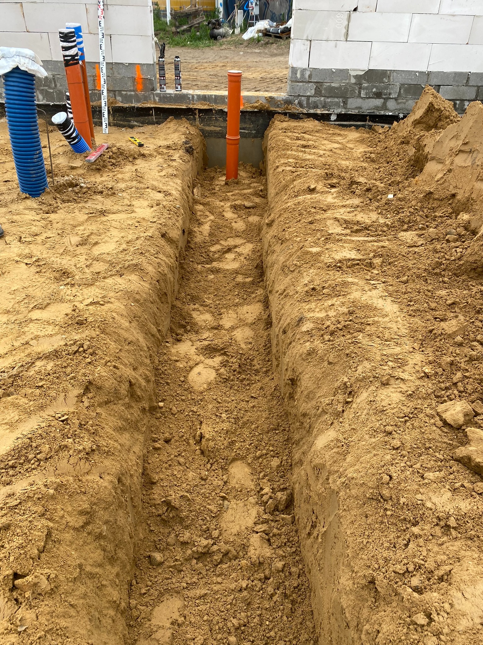 Wykop pod fundamenty z ułożonymi rurami wentylacyjnymi i instalacyjnymi, widoczne narzędzia pomiarowe i bloczki fundamentowe. Prace ziemne w toku.