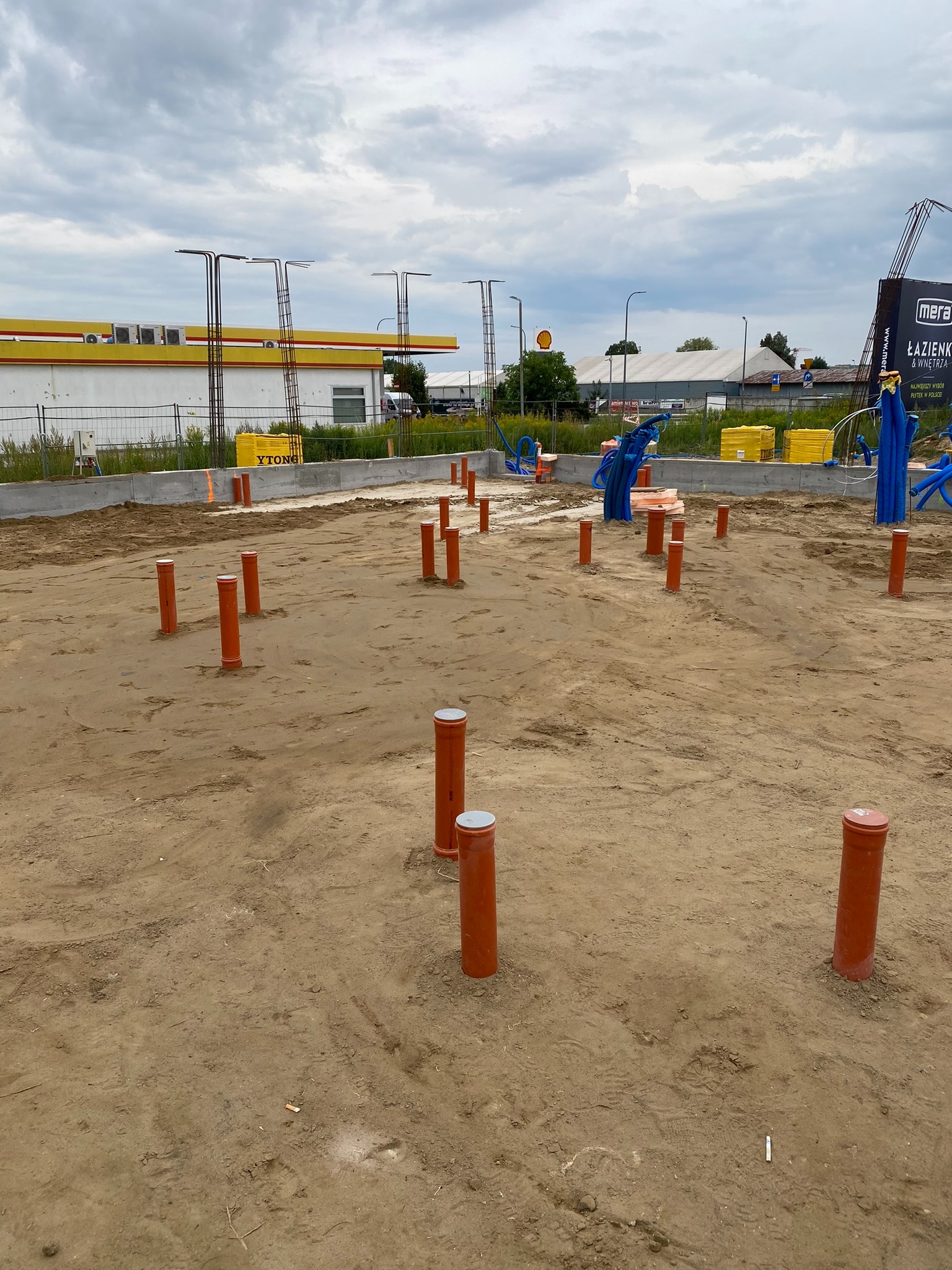 Fundamenty budynku z wystającymi rurami wentylacyjnymi, widoczne niebieskie rury i żółte bloczki Ytong. W tle stacja benzynowa Shell i reklama Mera Łazienki.