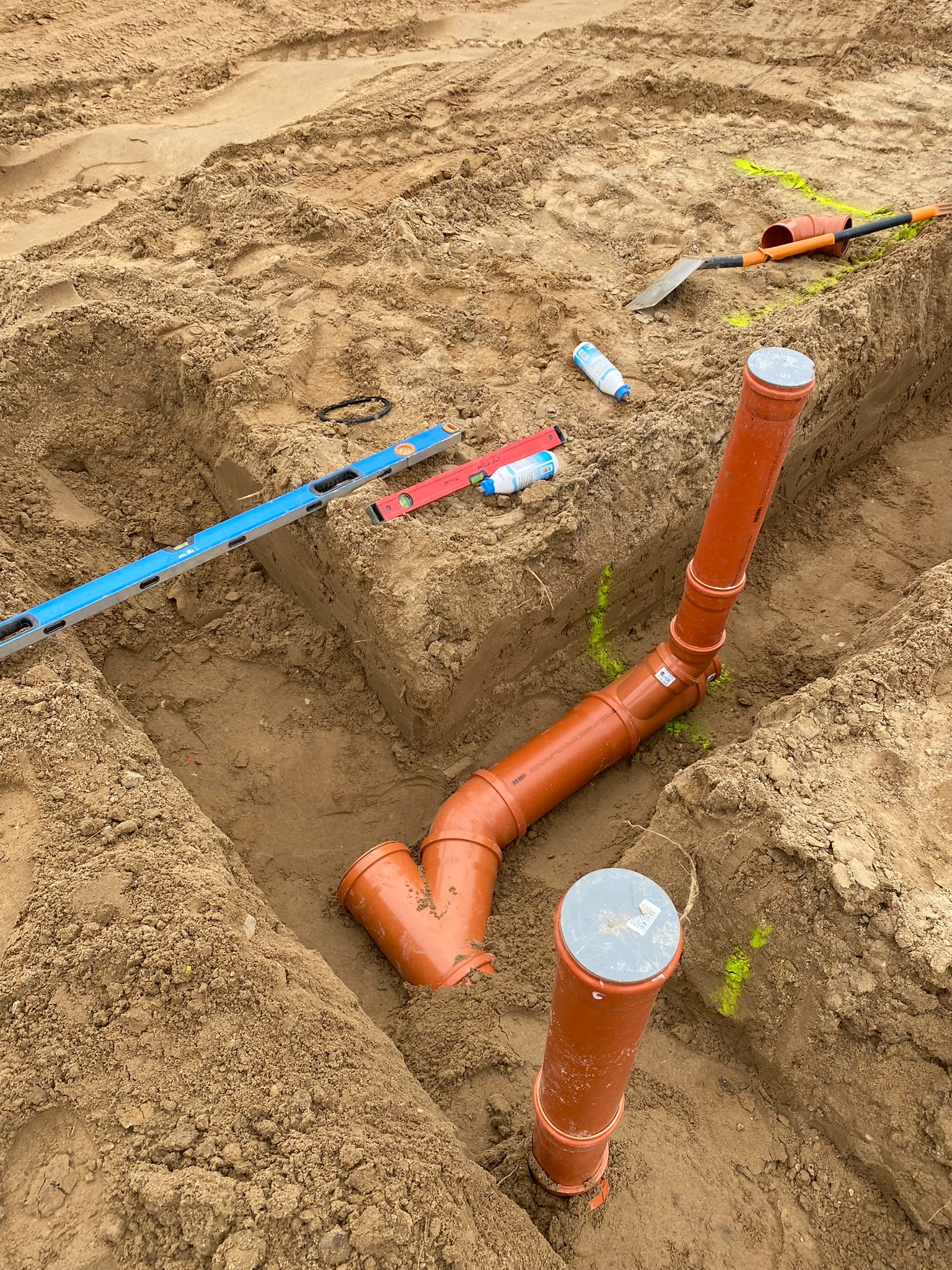 Montaż rur kanalizacyjnych w wykopie, widoczne poziomice i narzędzia. Żółte oznaczenia na ziemi. Prace instalacyjne w trakcie realizacji.