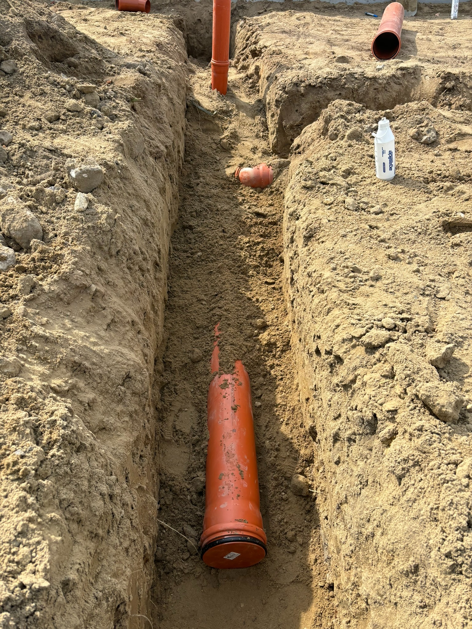 Wykop w ziemi z pomarańczowymi rurami kanalizacyjnymi w trakcie instalacji. Widoczna butelka środka smarnego do rur. Prace ziemne w słoneczny dzień.