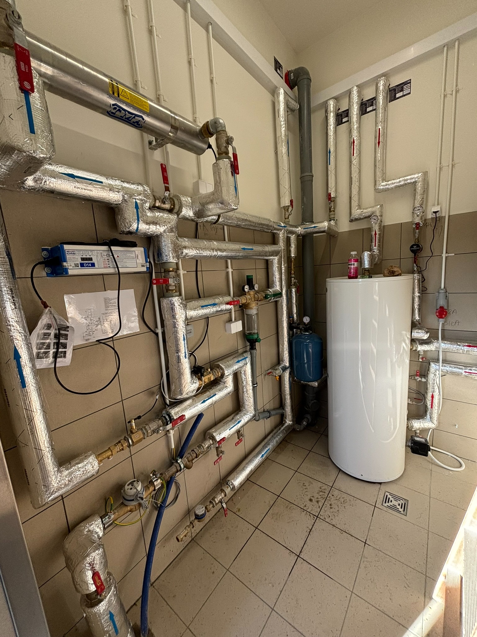 Złożona instalacja rur z izolacją termiczną, zaworami i elementami pomiarowymi w pomieszczeniu z wykończeniem ceramicznym. Widoczny biały zbiornik i niebieski element instalacji.
