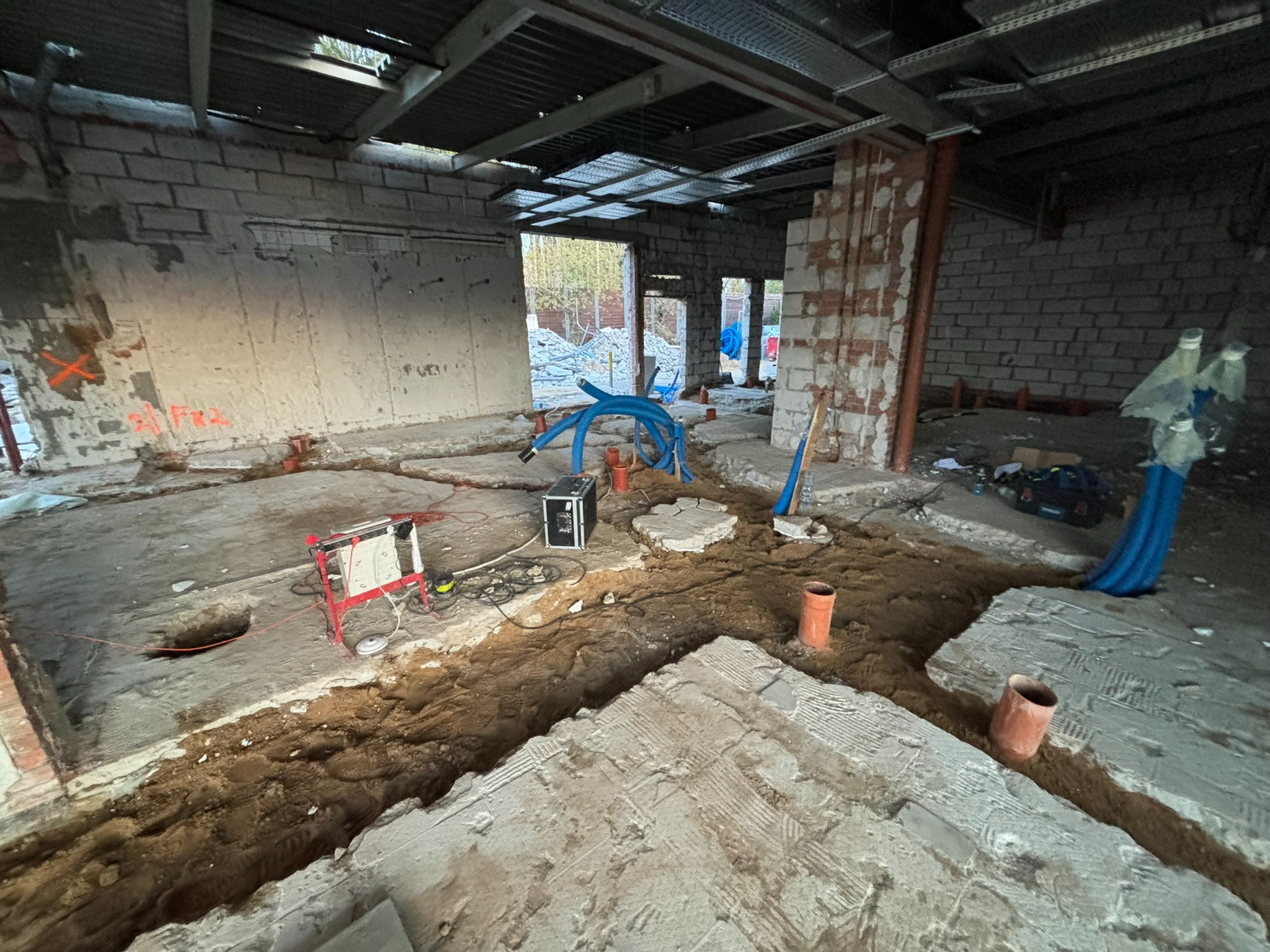 Wnętrze w trakcie prac instalacyjnych: wykopane rowy w podłodze, widoczne rury wentylacyjne, sprzęt pomiarowy. Surowy stan, cegły, beton, przygotowanie do dalszych etapów.