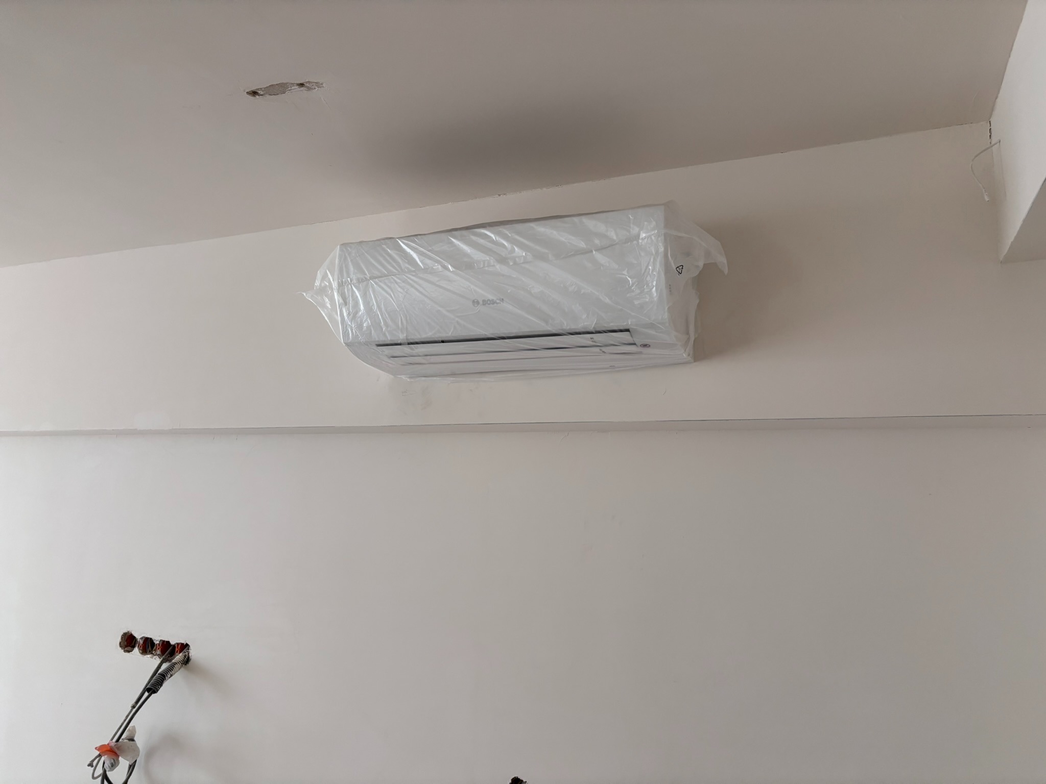 Biały klimatyzator Bosch w folii ochronnej, zamontowany na jasnej ścianie. Widoczne przygotowane otwory na kable i instalację elektryczną.