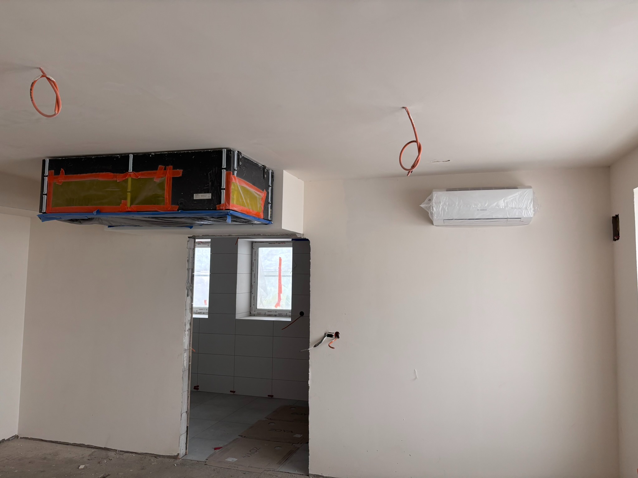 Montaż klimatyzacji kanałowej i ściennej w surowym wnętrzu. Widoczne zabezpieczenia folią i taśmą. Przewody elektryczne wystają ze ściany i sufitu.