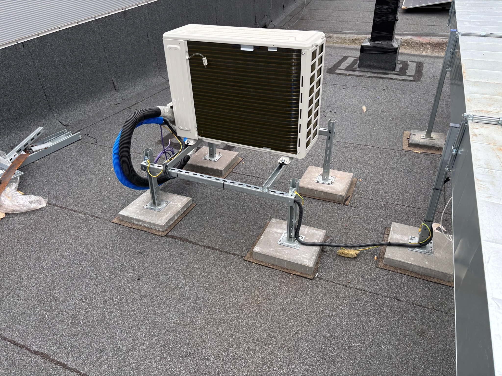 Zewnętrzna jednostka klimatyzacji na dachu, zamontowana na metalowej konstrukcji z betonowymi podstawami. Widoczne przewody i elementy instalacji.