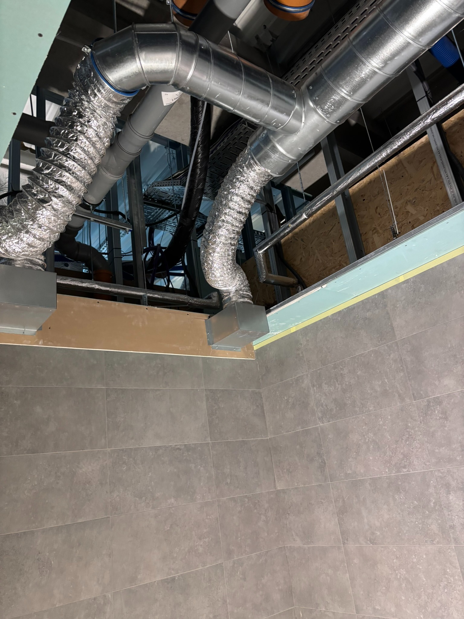 Srebrne, elastyczne rury wentylacyjne zamontowane pod sufitem podwieszanym z płyt gipsowo-kartonowych. Widoczne elementy konstrukcji i instalacji.