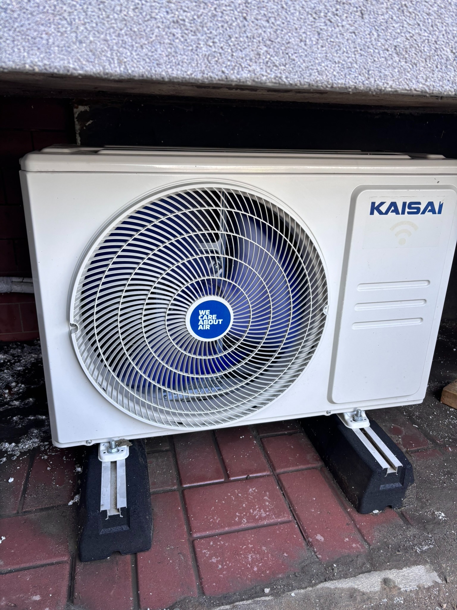 Biała jednostka zewnętrzna klimatyzacji Kaisai z niebieskim wentylatorem, zamontowana na czarnych wspornikach na czerwonej kostce brukowej, widok z dołu.