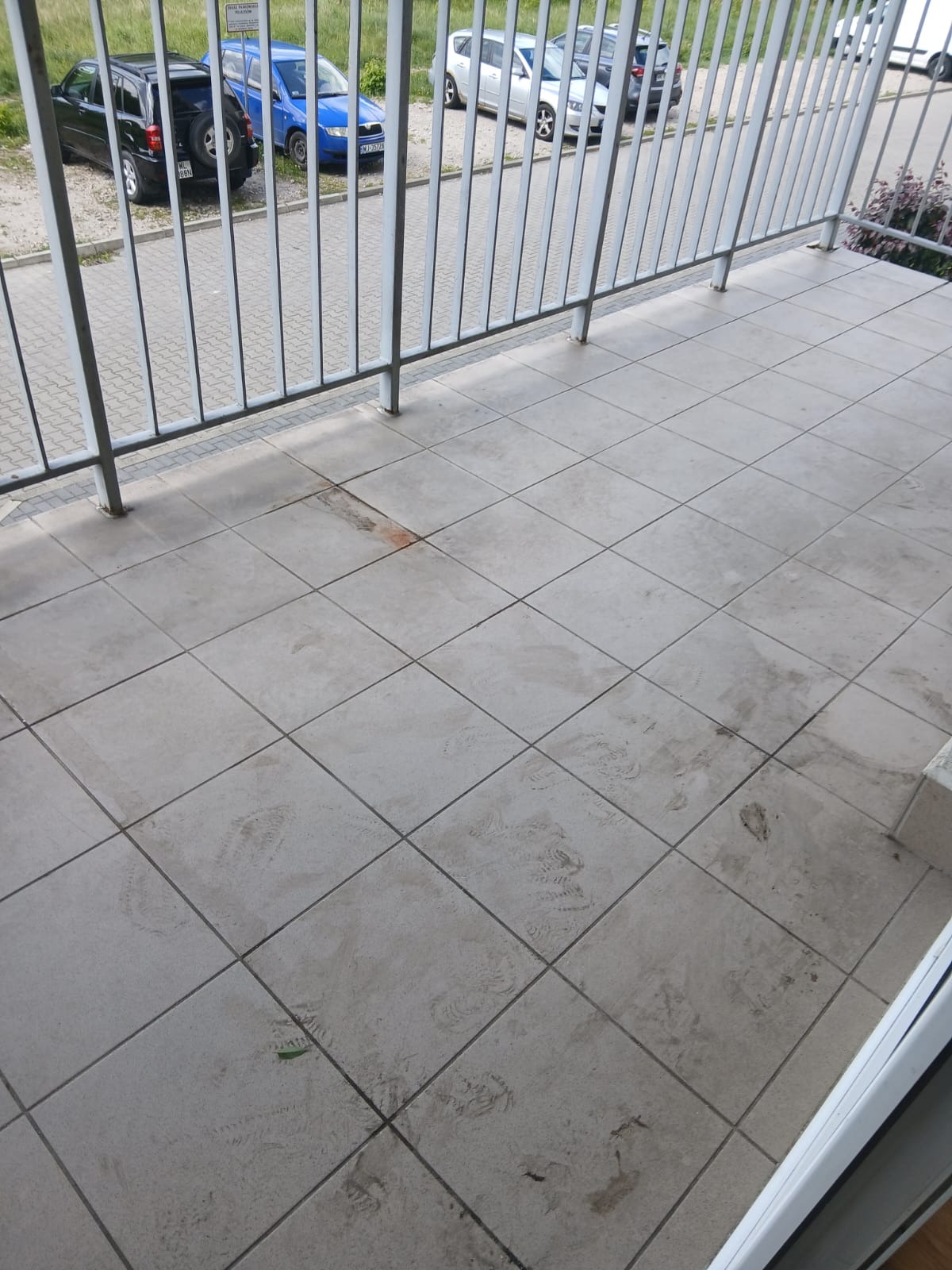 Brudna, wykafelkowana podłoga balkonu z widocznymi śladami stóp i zabrudzeniami. W tle barierka i zaparkowane samochody.