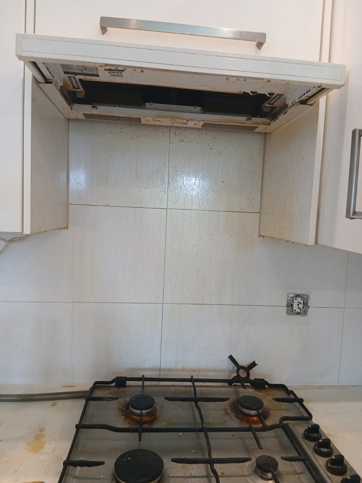 Zanieczyszczona kuchnia: tłusty okap, brudne płytki nad kuchenką gazową. Widoczny otwarty kontakt elektryczny i elementy armatury. Wymaga gruntownego sprzątania.