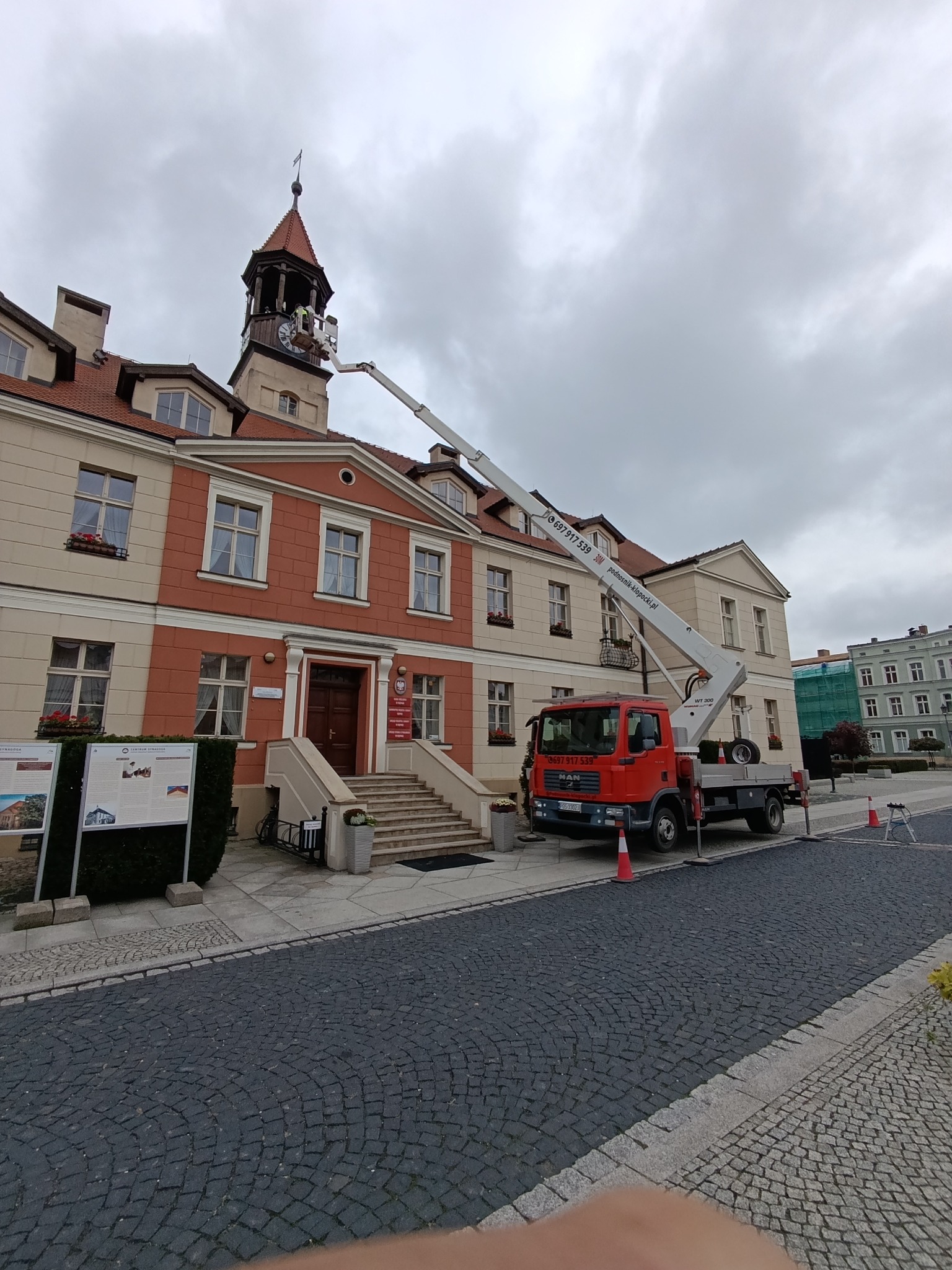 Czerwony podnośnik koszowy na placu, pracownik w koszu przy wieży zegarowej zabytkowego budynku. Usługa konserwacji zegara miejskiego w pochmurny dzień.