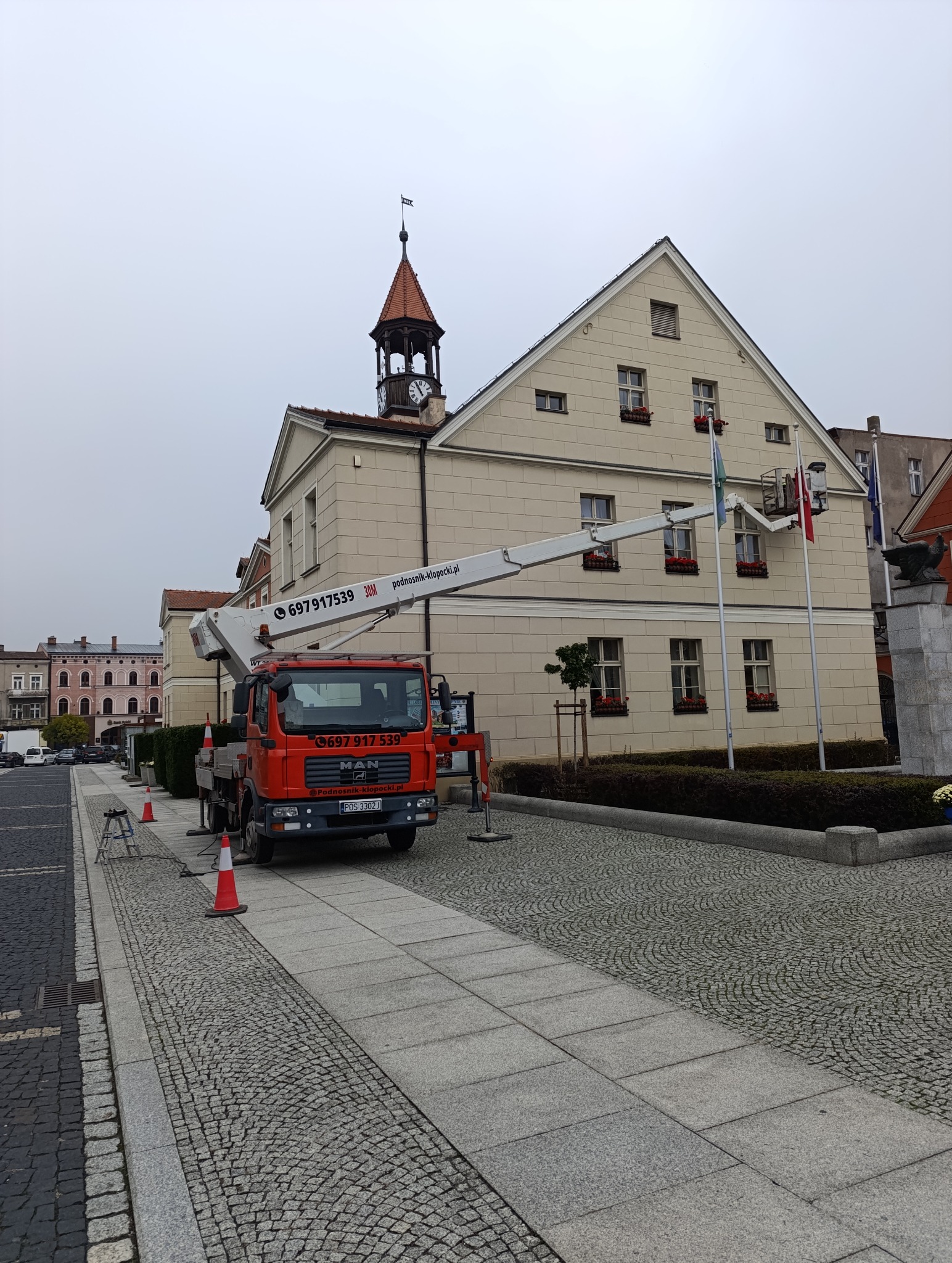 Czerwony podnośnik koszowy MAN na tle beżowego budynku z wieżą zegarową. Kosz podnośnika przy fladze. Brukowana ulica i sygnalizacyjne pachołki.