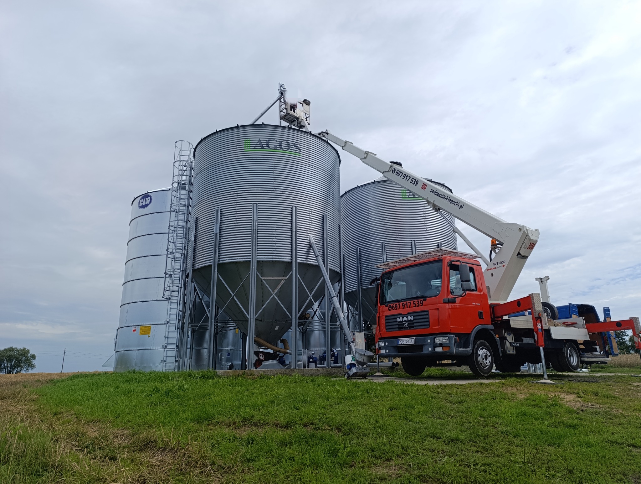 Podnośnik koszowy MAN z wysięgnikiem przy silosach zbożowych AGOS i CIN. Widoczne numery kontaktowe i adres strony internetowej na ramieniu podnośnika.