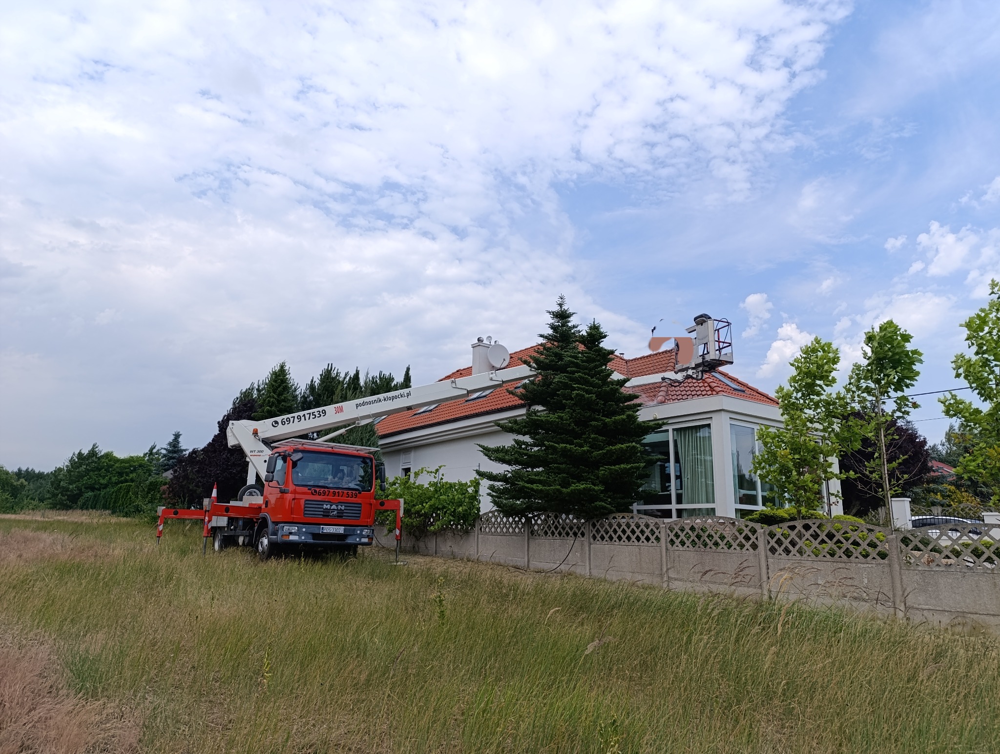 Czerwony podnośnik koszowy MAN na tle domu z czerwoną dachówką. Kosz podnośnika na wysokości dachu, sugeruje prace na wysokościach. Letni dzień, błękitne niebo.