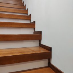 Stolarstwo Janicki Henryk - Detal brązowych, drewnianych schodów z widoczną strukturą drewna, na tle białej ściany. Minimalistyczny design.