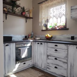 Stolarstwo Janicki Henryk - Rustykalna kuchnia z białymi, postarzanymi frontami. Blat w kolorze ciemnego grafitu. Na blacie owoce, przyprawy i dekoracje. Okno zdobi koronkowa firanka.